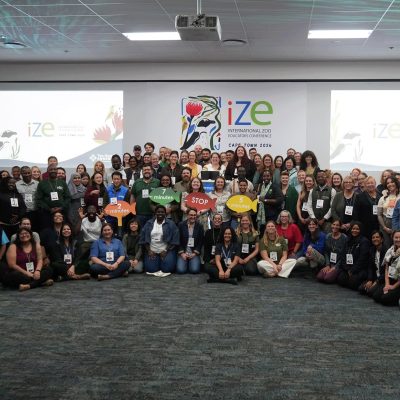 A large group of conference delegates stands and crouches together for a group photograph in front of a screen reading IZE International Zoo Educators Conference Cape Town 2026.