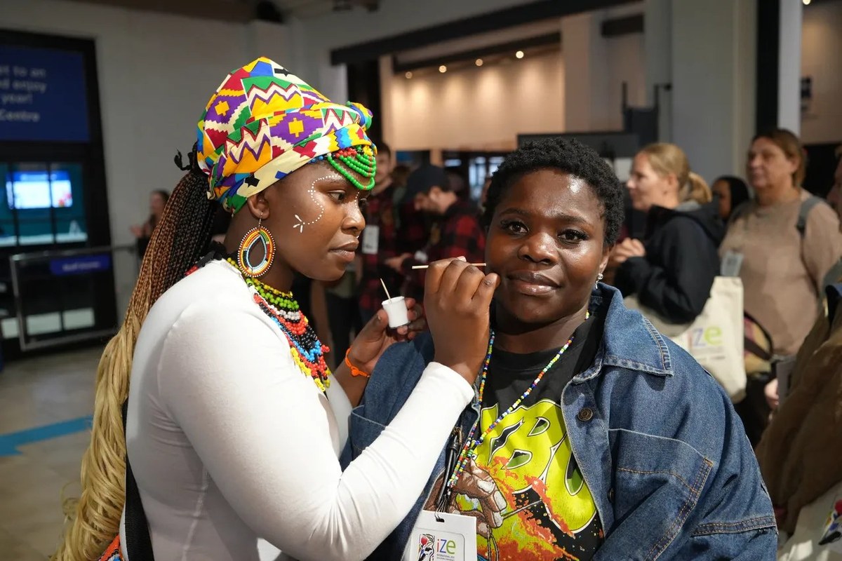 A woman in a brightly patterned headwrap and beaded jewelry paints a design on the face of a conference delegate wearing an IZE lanyard at the Two Oceans Aquarium