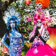 Two performers dressed as La Catrina at the Houston Zoo, in full calavera face paint and towering floral headdresses, one in blue and white patterned dress and one in pink, standing under oak trees hung with orange and red lanterns.