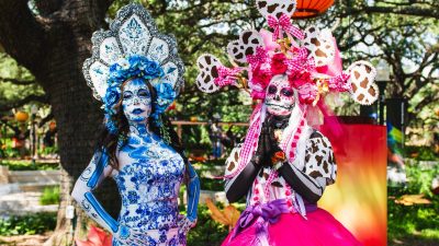Two performers dressed as La Catrina at the Houston Zoo, in full calavera face paint and towering floral headdresses, one in blue and white patterned dress and one in pink, standing under oak trees hung with orange and red lanterns.