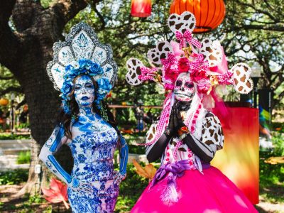 Two performers dressed as La Catrina at the Houston Zoo, in full calavera face paint and towering floral headdresses, one in blue and white patterned dress and one in pink, standing under oak trees hung with orange and red lanterns.