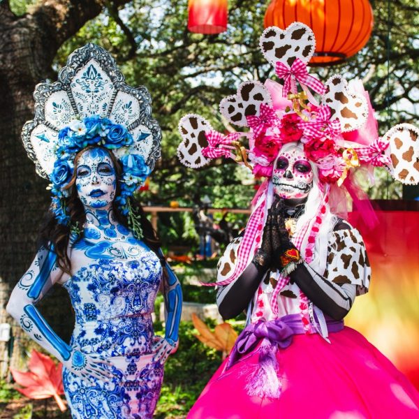 Two performers dressed as La Catrina at the Houston Zoo, in full calavera face paint and towering floral headdresses, one in blue and white patterned dress and one in pink, standing under oak trees hung with orange and red lanterns.