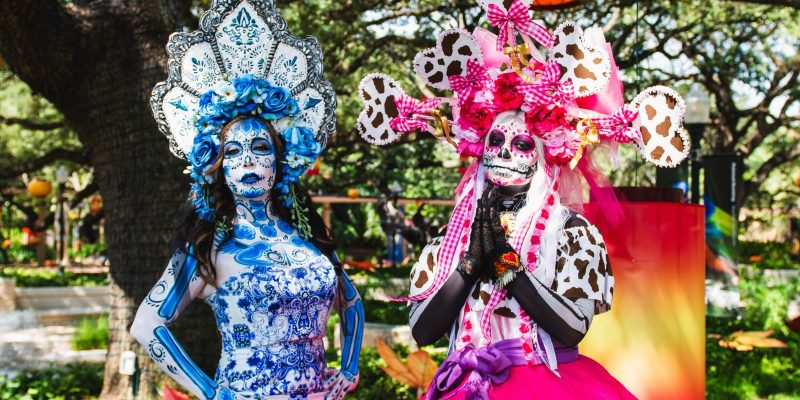 Two performers dressed as La Catrina at the Houston Zoo, in full calavera face paint and towering floral headdresses, one in blue and white patterned dress and one in pink, standing under oak trees hung with orange and red lanterns.