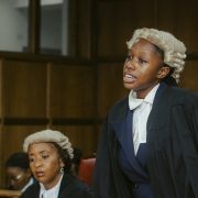 A law student in a barrister's wig and gown stands to address the court while a judge listens from the side.