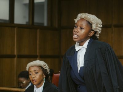 A law student in a barrister's wig and gown stands to address the court while a judge listens from the side.