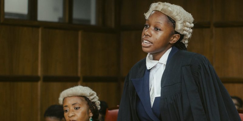 A law student in a barrister's wig and gown stands to address the court while a judge listens from the side.