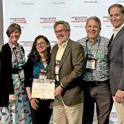 Six people stand together in front of a backdrop printed with the Association of Zoos and Aquariums logo, two of them holding a framed award certificate between them.
