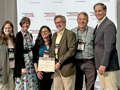 Six people stand together in front of a backdrop printed with the Association of Zoos and Aquariums logo, two of them holding a framed award certificate between them.