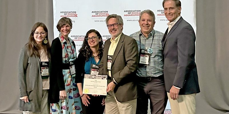 Six people stand together in front of a backdrop printed with the Association of Zoos and Aquariums logo, two of them holding a framed award certificate between them.