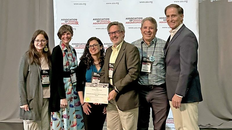 Six people stand together in front of a backdrop printed with the Association of Zoos and Aquariums logo, two of them holding a framed award certificate between them.