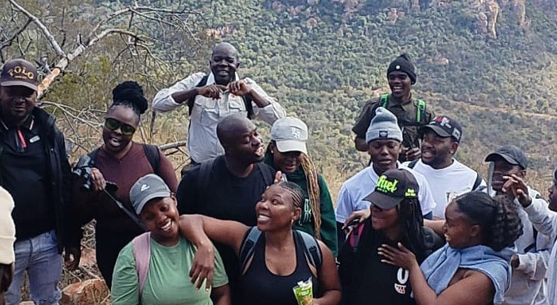 A large group of young hikers in caps and beanies standing close together and laughing on a rocky outcrop at Medike Nature Reserve, with the wooded slopes of the Soutpansberg rising behind them.