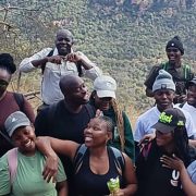 A large group of young hikers in caps and beanies standing close together and laughing on a rocky outcrop at Medike Nature Reserve, with the wooded slopes of the Soutpansberg rising behind them.