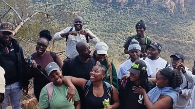 A large group of young hikers in caps and beanies standing close together and laughing on a rocky outcrop at Medike Nature Reserve, with the wooded slopes of the Soutpansberg rising behind them.