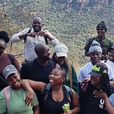 A large group of young hikers in caps and beanies standing close together and laughing on a rocky outcrop at Medike Nature Reserve, with the wooded slopes of the Soutpansberg rising behind them.