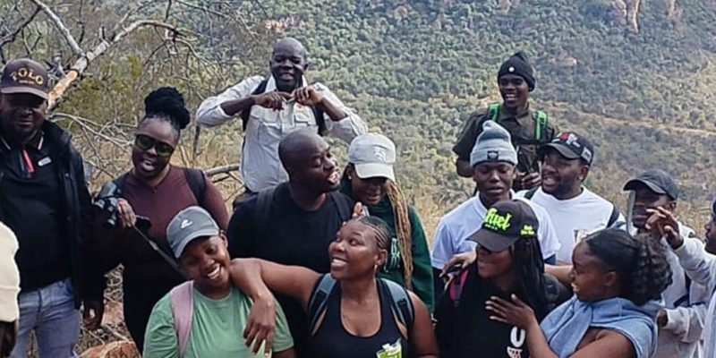 A large group of young hikers in caps and beanies standing close together and laughing on a rocky outcrop at Medike Nature Reserve, with the wooded slopes of the Soutpansberg rising behind them.