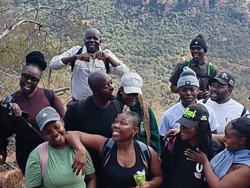 A large group of young hikers in caps and beanies standing close together and laughing on a rocky outcrop at Medike Nature Reserve, with the wooded slopes of the Soutpansberg rising behind them.