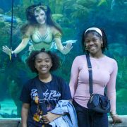 Two young guests smile for a photograph at the glass of the Great Maya Reef exhibit at Audubon Aquarium while a mermaid performer in a gold and green tail hovers in the water behind them with her arms outstretched.