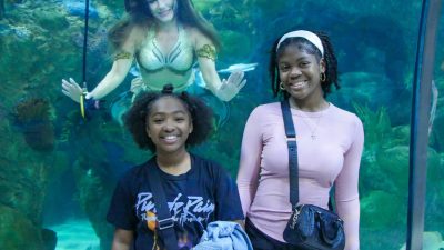 Two young guests smile for a photograph at the glass of the Great Maya Reef exhibit at Audubon Aquarium while a mermaid performer in a gold and green tail hovers in the water behind them with her arms outstretched.