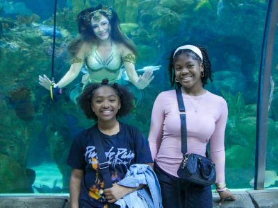 Two young guests smile for a photograph at the glass of the Great Maya Reef exhibit at Audubon Aquarium while a mermaid performer in a gold and green tail hovers in the water behind them with her arms outstretched.