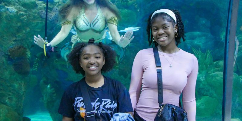 Two young guests smile for a photograph at the glass of the Great Maya Reef exhibit at Audubon Aquarium while a mermaid performer in a gold and green tail hovers in the water behind them with her arms outstretched.