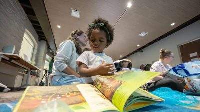 A young child in a white T-shirt leans over a large open picture book on a table, an adult program leader crouched beside them, with other families reading on the floor of the room behind.