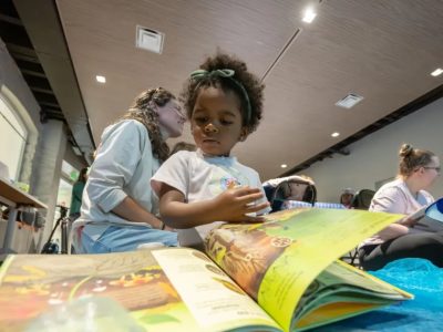 A young child in a white T-shirt leans over a large open picture book on a table, an adult program leader crouched beside them, with other families reading on the floor of the room behind.