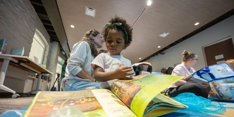 A young child in a white T-shirt leans over a large open picture book on a table, an adult program leader crouched beside them, with other families reading on the floor of the room behind.