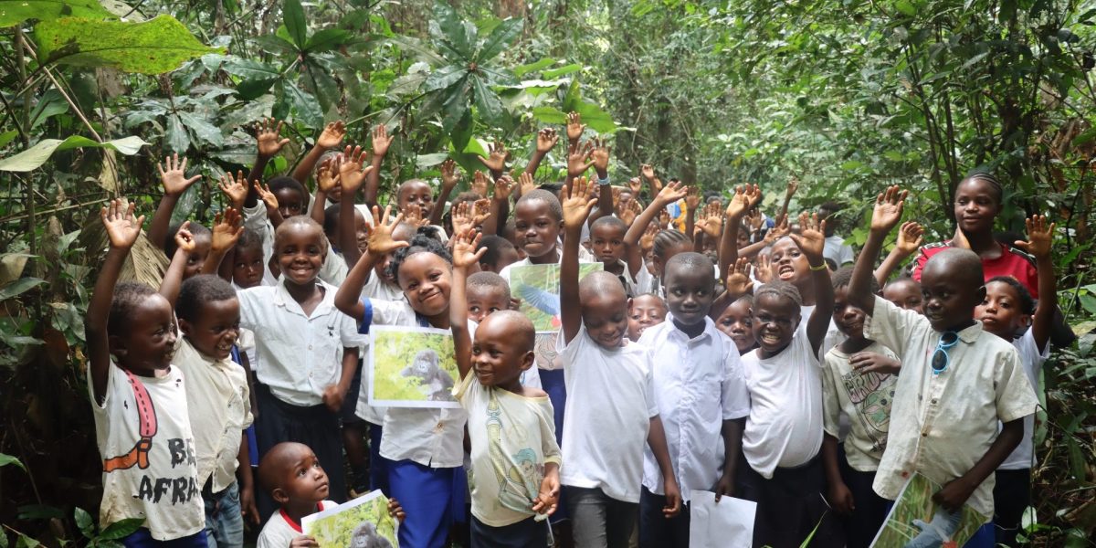 Dozens of children stand among broad-leaved forest plants near Nkuba with their hands raised, several holding printed photographs of gorillas and birds.