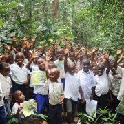 Dozens of children stand among broad-leaved forest plants near Nkuba with their hands raised, several holding printed photographs of gorillas and birds.