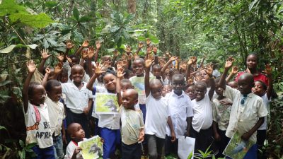 Dozens of children stand among broad-leaved forest plants near Nkuba with their hands raised, several holding printed photographs of gorillas and birds.