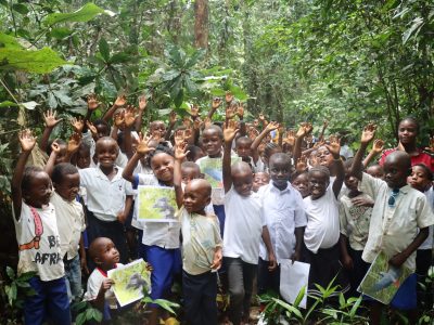 Dozens of children stand among broad-leaved forest plants near Nkuba with their hands raised, several holding printed photographs of gorillas and birds.