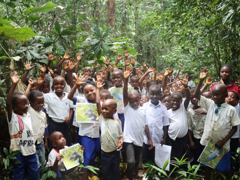 Dozens of children stand among broad-leaved forest plants near Nkuba with their hands raised, several holding printed photographs of gorillas and birds.