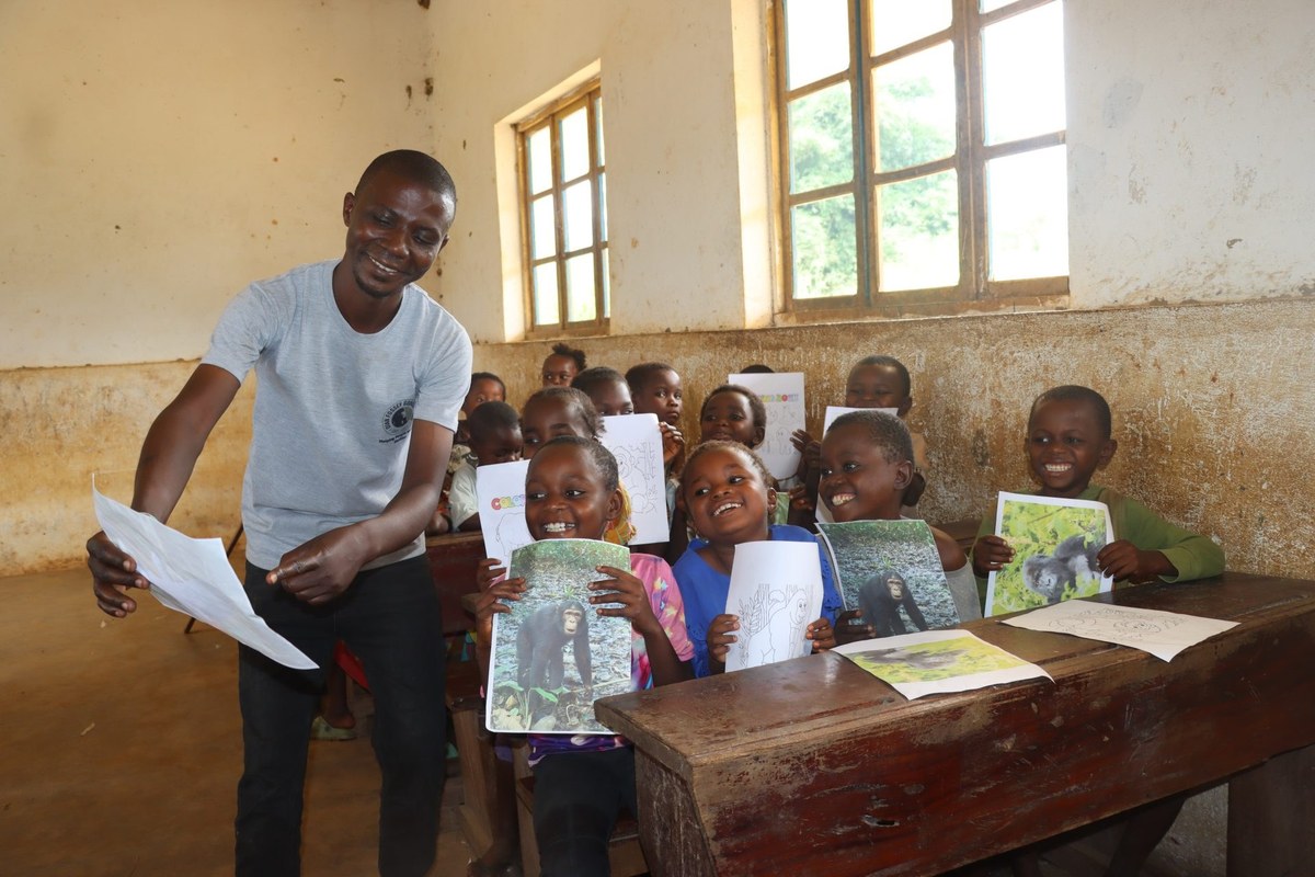 A Dian Fossey Gorilla Fund teacher hands out worksheets in a bare classroom while children at a wooden desk hold up photographs of gorillas and coloring sheets