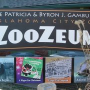 The entrance sign for the Patricia and Byron J. Gambulos ZooZeum at the Oklahoma City Zoo, a large dark sign in white and orange lettering mounted above four framed panels on the zoo's history, with dry ornamental grass in the foreground.