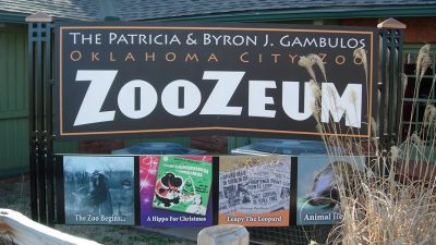 The entrance sign for the Patricia and Byron J. Gambulos ZooZeum at the Oklahoma City Zoo, a large dark sign in white and orange lettering mounted above four framed panels on the zoo's history, with dry ornamental grass in the foreground.