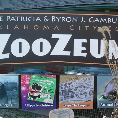 The entrance sign for the Patricia and Byron J. Gambulos ZooZeum at the Oklahoma City Zoo, a large dark sign in white and orange lettering mounted above four framed panels on the zoo's history, with dry ornamental grass in the foreground.