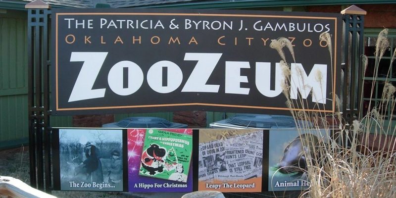 The entrance sign for the Patricia and Byron J. Gambulos ZooZeum at the Oklahoma City Zoo, a large dark sign in white and orange lettering mounted above four framed panels on the zoo's history, with dry ornamental grass in the foreground.