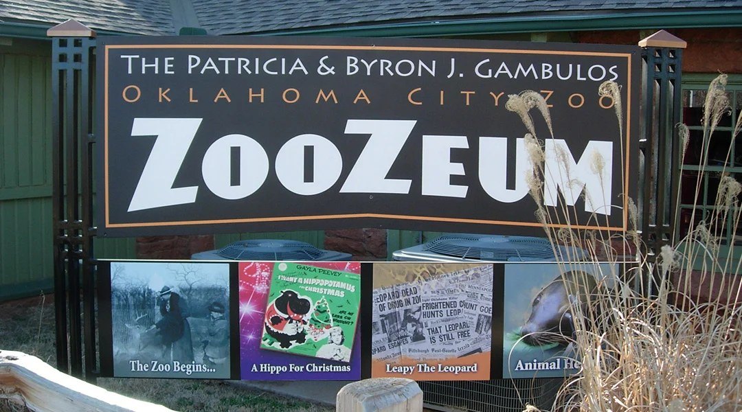 The entrance sign for the Patricia and Byron J. Gambulos ZooZeum at the Oklahoma City Zoo, a large dark sign in white and orange lettering mounted above four framed panels on the zoo's history, with dry ornamental grass in the foreground.
