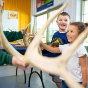 Two young children in Lee G. Simmons Wildlife Safari Park shirts smile as a staff member in a green uniform holds up a large branching antler in a bright indoor education room, with more antlers and animal skulls laid out on a table behind them.