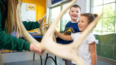 Two young children in Lee G. Simmons Wildlife Safari Park shirts smile as a staff member in a green uniform holds up a large branching antler in a bright indoor education room, with more antlers and animal skulls laid out on a table behind them.