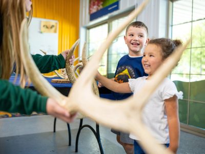 Two young children in Lee G. Simmons Wildlife Safari Park shirts smile as a staff member in a green uniform holds up a large branching antler in a bright indoor education room, with more antlers and animal skulls laid out on a table behind them.