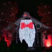 A giant illuminated penguin sculpture wearing a red bow tie towers over visitors in silhouette at night during LumiNature at Philadelphia Zoo, with red-lit trees and a building behind it.