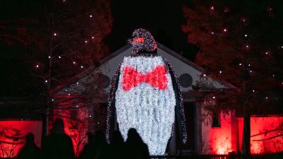 A giant illuminated penguin sculpture wearing a red bow tie towers over visitors in silhouette at night during LumiNature at Philadelphia Zoo, with red-lit trees and a building behind it.