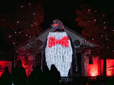A giant illuminated penguin sculpture wearing a red bow tie towers over visitors in silhouette at night during LumiNature at Philadelphia Zoo, with red-lit trees and a building behind it.
