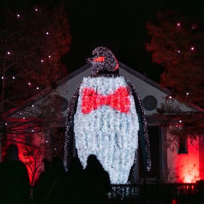 A giant illuminated penguin sculpture wearing a red bow tie towers over visitors in silhouette at night during LumiNature at Philadelphia Zoo, with red-lit trees and a building behind it.
