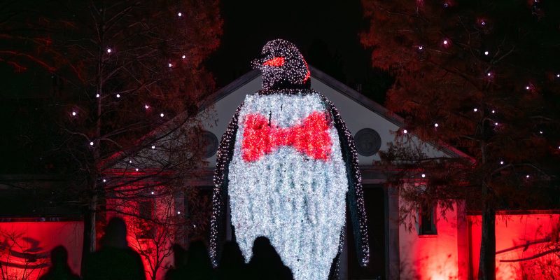 A giant illuminated penguin sculpture wearing a red bow tie towers over visitors in silhouette at night during LumiNature at Philadelphia Zoo, with red-lit trees and a building behind it.