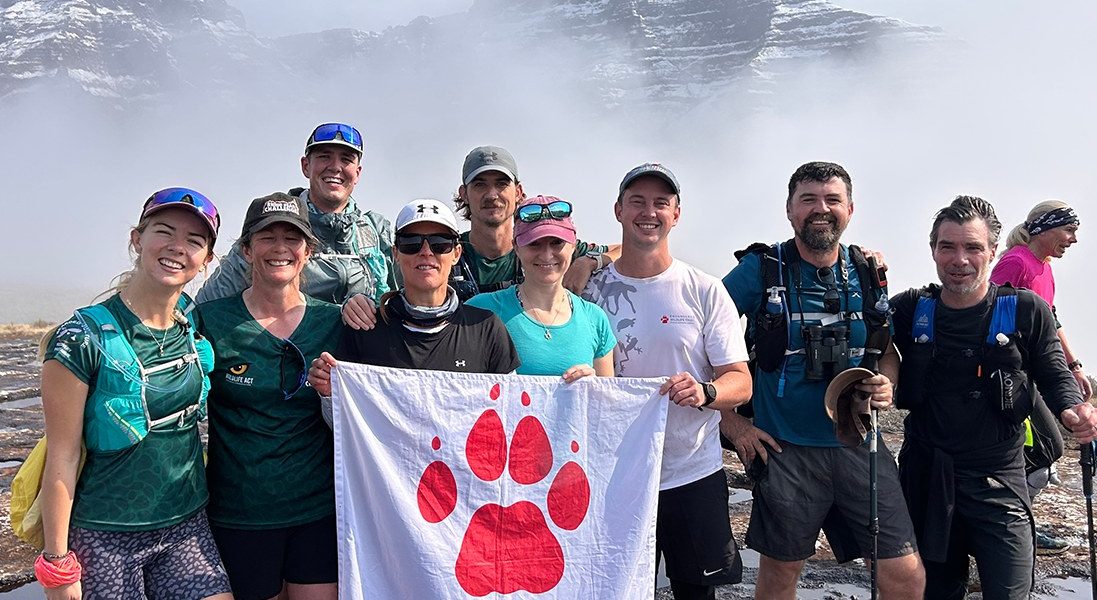 Ten hikers in trail running kit stand together on wet rock holding a white flag with a red paw print reading Endangered Wildlife Trust, with a snow covered Drakensberg escarpment rising out of thick cloud behind them.