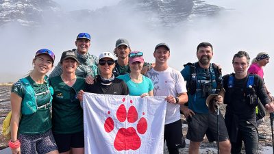 Ten hikers in trail running kit stand together on wet rock holding a white flag with a red paw print reading Endangered Wildlife Trust, with a snow covered Drakensberg escarpment rising out of thick cloud behind them.