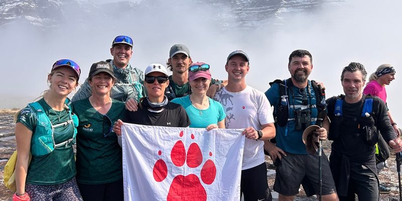 Ten hikers in trail running kit stand together on wet rock holding a white flag with a red paw print reading Endangered Wildlife Trust, with a snow covered Drakensberg escarpment rising out of thick cloud behind them.