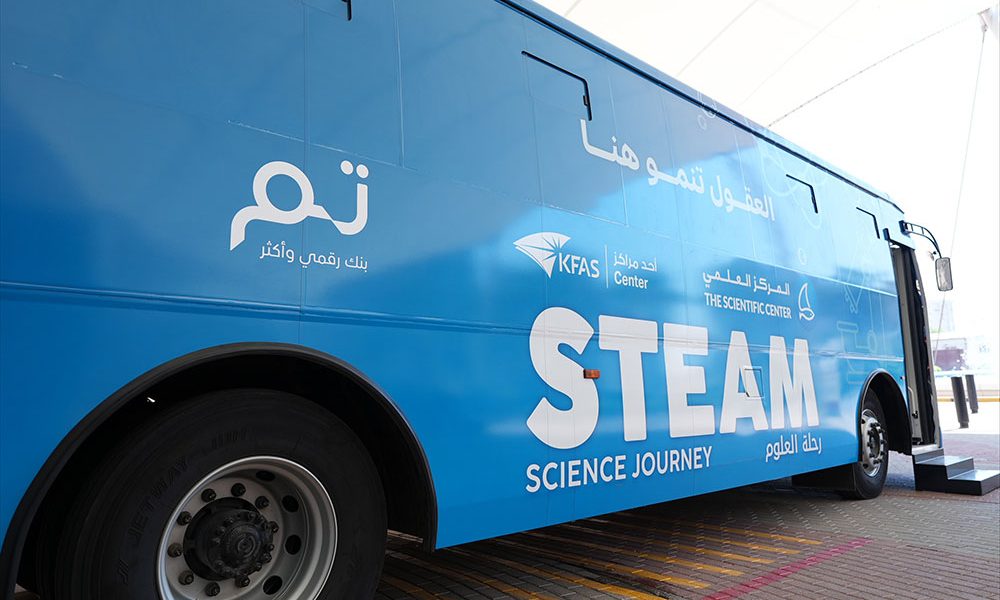 A blue coach parked on paving with its door open, lettered STEAM Science Journey in English and Arabic alongside the logos of KFAS, The Scientific Center and Tam.